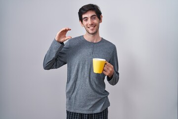 Poster - Young hispanic man wearing pajama drinking a cup of coffee smiling and confident gesturing with hand doing small size sign with fingers looking and the camera. measure concept.