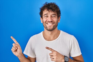 Poster - Hispanic young man standing over blue background smiling and looking at the camera pointing with two hands and fingers to the side.