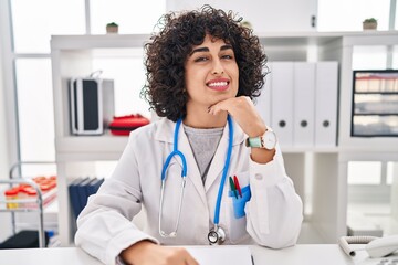 Sticker - Young brunette woman with curly hair wearing doctor uniform and stethoscope smiling looking confident at the camera with crossed arms and hand on chin. thinking positive.