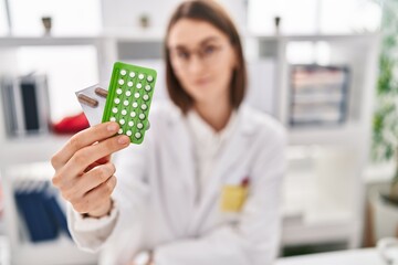 Poster - Young caucasian woman doctor holding birth control pills sitting on table at clinic