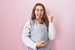 Young caucasian woman holding and carrying baby on a sling screaming proud, celebrating victory and success very excited with raised arms