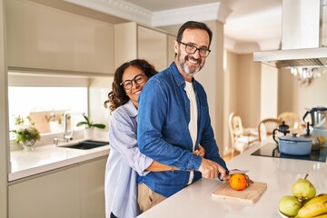 Wall Mural - Middle age hispanic couple hugging each other cutting orange at kitchen