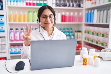 Wall Mural - Young arab woman working at pharmacy drugstore using laptop showing and pointing up with fingers number three while smiling confident and happy.