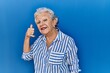 © Krakenimages.com - Senior woman with grey hair standing over blue background smiling doing phone gesture with hand and fingers like talking on the telephone. communicating concepts.