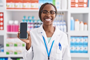 Canvas Print - African american woman working at pharmacy drugstore showing smartphone screen looking positive and happy standing and smiling with a confident smile showing teeth