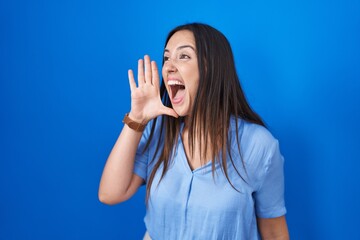 Sticker - Young brunette woman standing over blue background shouting and screaming loud to side with hand on mouth. communication concept.