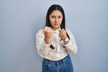 Sticker - Young latin woman standing over blue background ready to fight with fist defense gesture, angry and upset face, afraid of problem