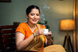 © WESTOCK - Happy smiling woman drinking tea or coffee by looking at camera while sitting on chair at home - concept of mourning routine, healthy diet and satisfaction