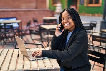 Wall Mural - African american woman talking on smartphone using laptop sitting on table at coffee shop terrace