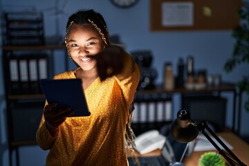 Canvas Print - African american woman with braids working at the office at night with tablet pointing to you and the camera with fingers, smiling positive and cheerful