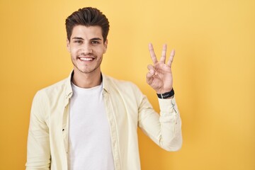 Sticker - Young hispanic man standing over yellow background showing and pointing up with fingers number three while smiling confident and happy.