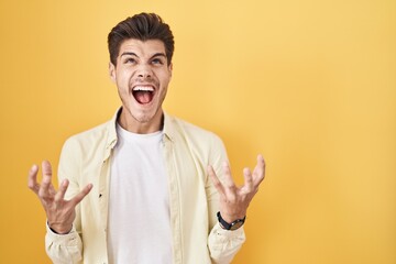 Sticker - Young hispanic man standing over yellow background crazy and mad shouting and yelling with aggressive expression and arms raised. frustration concept.