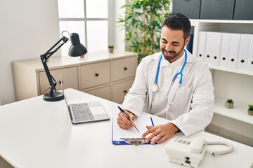 Wall Mural - Young hispanic man wearing doctor uniform writing on clipboard at clinic