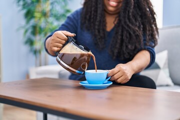 Canvas Print - African american woman pouring coffee on cup sitting on sofa at home