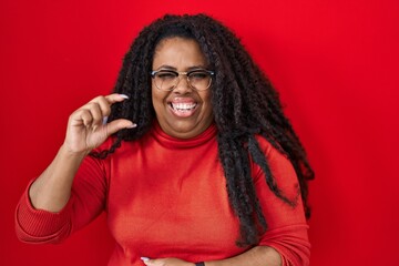 Sticker - Plus size hispanic woman standing over red background smiling and confident gesturing with hand doing small size sign with fingers looking and the camera. measure concept.