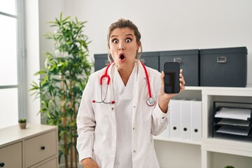 Poster - Young woman wearing doctor uniform holding smartphone scared and amazed with open mouth for surprise, disbelief face