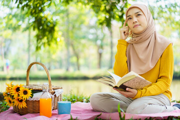 Wall Mural - Happy cheerful Asian beautiful muslim woman sitting on the ground at the park and drinking a cup of tea in afternoon. Beautiful muslim woman relaxing and picnic in weekend.