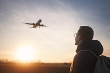 Man With Backpack Looking Up To Airplane Landing At Airport During Beautiful Sunset. .