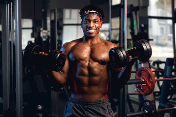 20s black and muscular man in a gym showing dumbbells and his muscles.
