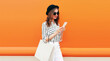 Portrait of beautiful young woman looking at smartphone with shopping bag wearing white striped shirt, black round hat on orange background