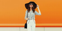 Portrait Of Beautiful Woman Model Wearing Black Round Straw Summer Hat, Handbag And White Striped Shirt On Orange Background