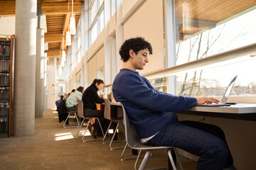 latin american student sitting at desk with laptop, preparing for examinations in the library campus