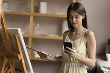 Positive Beautiful Young Artist Apprentice Using Smartphone At Easel, Consulting Internet For Creative Ideas, Chatting With Client, Customer, Reading Message, Holding Palette