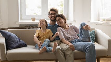 Happy handsome loving caring husband hugging cute laughing daughter girl and cheerful pretty wife, resting on home couch, relaxing in living room, enjoying family leisure time