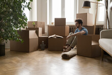 Young Property Buyer Man Sitting On Parqueted Heating Floor At Stacks Of Cardboard Relocation Boxes, Packing Stuff, Unpacking Containers, Moving Into New Apartment Flat