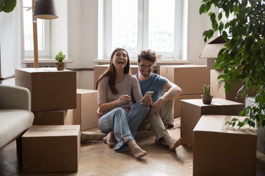 cheerful young couple of new homeowners sitting on floor at stacked moving boxes, using online app o