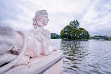 Ancient Park Sculpture - The Sphinx On The Background Of The Lake. Island With Trees On The Lake In Moscow Park.