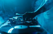 Night Club DJ Plays Set With Vinyl Records. Close Up Photo Of Professional Disc Jockey Mixing Music With Turntables And Sound Mixer In Smoke And Blue Lights