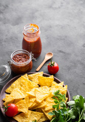 Wall Mural - Nachos or tortilla with spicy tomato sauce on a plate with fresh vegetables and herbs on a dark background. Corn chips with salsa sauce. Classic Mexican snack.