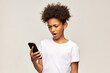 © Anatoliy Karlyuk - Perplexed upset afro teen boy in white mockup t-shirt standing against gray background with smartphone in hands, saying oh no, annoyed with running out battery. Children and negative emotions