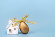 Happy Easter concept. One golden egg with a ribbon bow and white ceramic house on blue. Copy space