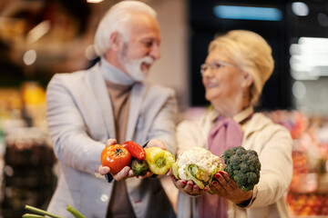 Wall Mural - Close up of senior couple showing fresh vegetables at the camera while standing at the hypermarket.