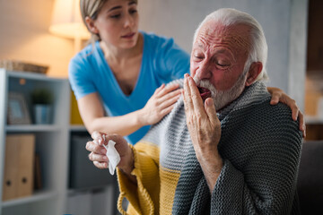 Wall Mural - Nice senior bearded man taking a paper tissue, sick at home. Female caregiver helping senior man. Female professional doctor touching shoulder comforting upset senior patient.