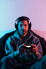 Young smiling male gamer wearing headset with mic and using controller while sitting on pouf playing video game