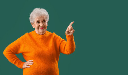 Wall Mural - Senior woman in orange sweater pointing at something on green background