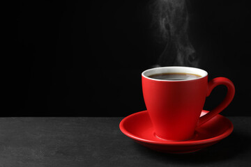 Wall Mural - Red cup with hot steaming coffee on grey table against black background, space for text