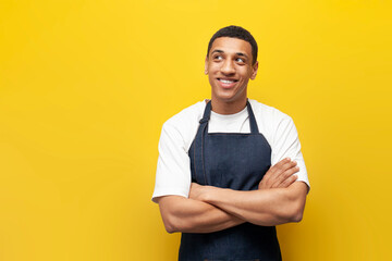 young guy afro american waiter in apron stands with his arms crossed on yellow isolated background, barista worker