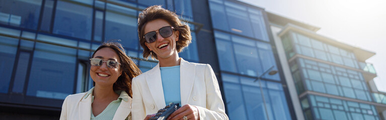 Two smiling woman in trendy outfits laugh and walk outside on background of modern building