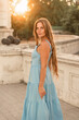 © svetograph - Woman sunset blue dress. Portrait of a woman with long hair and a blue dress against the backdrop of the setting sun and a white building. Lifestyle, walking around the city