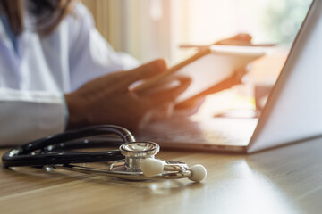 Wall Mural - Female doctor in white coat, hand holding and using modern digital tablet, work on laptop computer with stethoscope on the desk at office. Online medical, e health or medical network concept. 