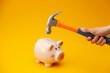 A pink piggy bank is about to be hit by a hammer. A hand holding a hammer which is raised above a pink china piggy bank, with a shocked and apprehensive facial expression. Financial problem.