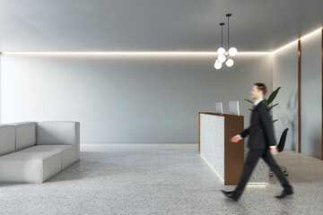 Poster - Businessman walking by modern reception desk in spacious office area with cozy grey sofa, concrete floor and light grey wall background