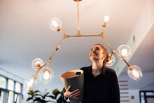 Happy Businesswoman Standing With Garbage Bin Below Illuminated Lighting Equipment In Office