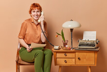 Positive Red Head Retro Businesswoman Works In Own Cabinet From Home Holds Paper Document Uses Typewriter Has Telephone Conversation Wears Shirt And Trousers Sits On Chair. Vintage Furniture