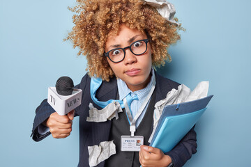 Shocked woman with curly hair stares at camera wears spectacles and formal outfit holds folders and microphone prepares for taking interview or giving speech during live stream. Mass media concept