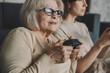 Cute emotional senior woman and daughter playing videogame holding gamepad controllers sitting on sofa at home. Having fun on weekend. Family of two having fun